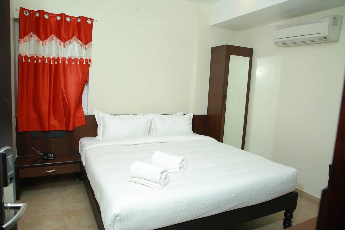 A hotel room with a bed, white linens, a red curtain, a wooden wardrobe, and an air conditioning unit.