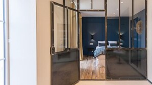 A bedroom with a large sliding glass door leading to a balcony, a bed with a blue bedspread, a bedside table with a lamp, and a blue wall. - Maison Deveney Mars (Puligny-Montrachet)