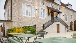 A stone building with a staircase leading to a wooden door. There are outdoor tables and chairs. - Maison Deveney Mars (Puligny-Montrachet)