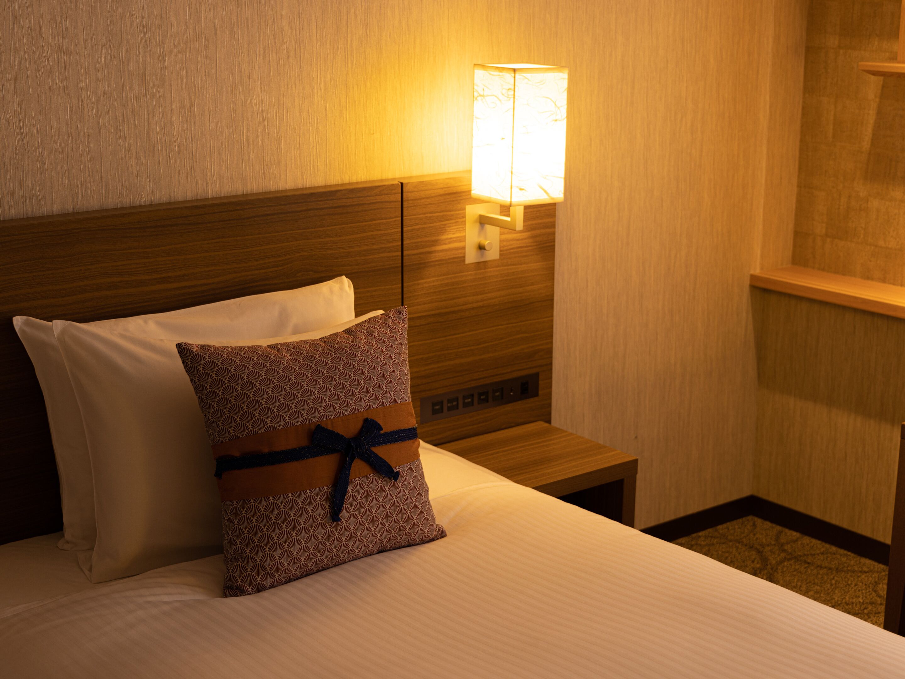 A hotel room with a neatly made bed, a nightstand with a lamp, and a wooden headboard.