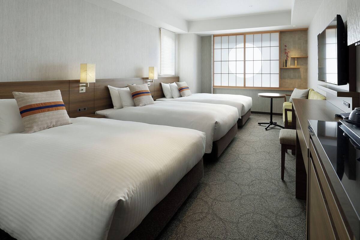 A hotel room with three beds, a desk, a television, and a window with blinds.