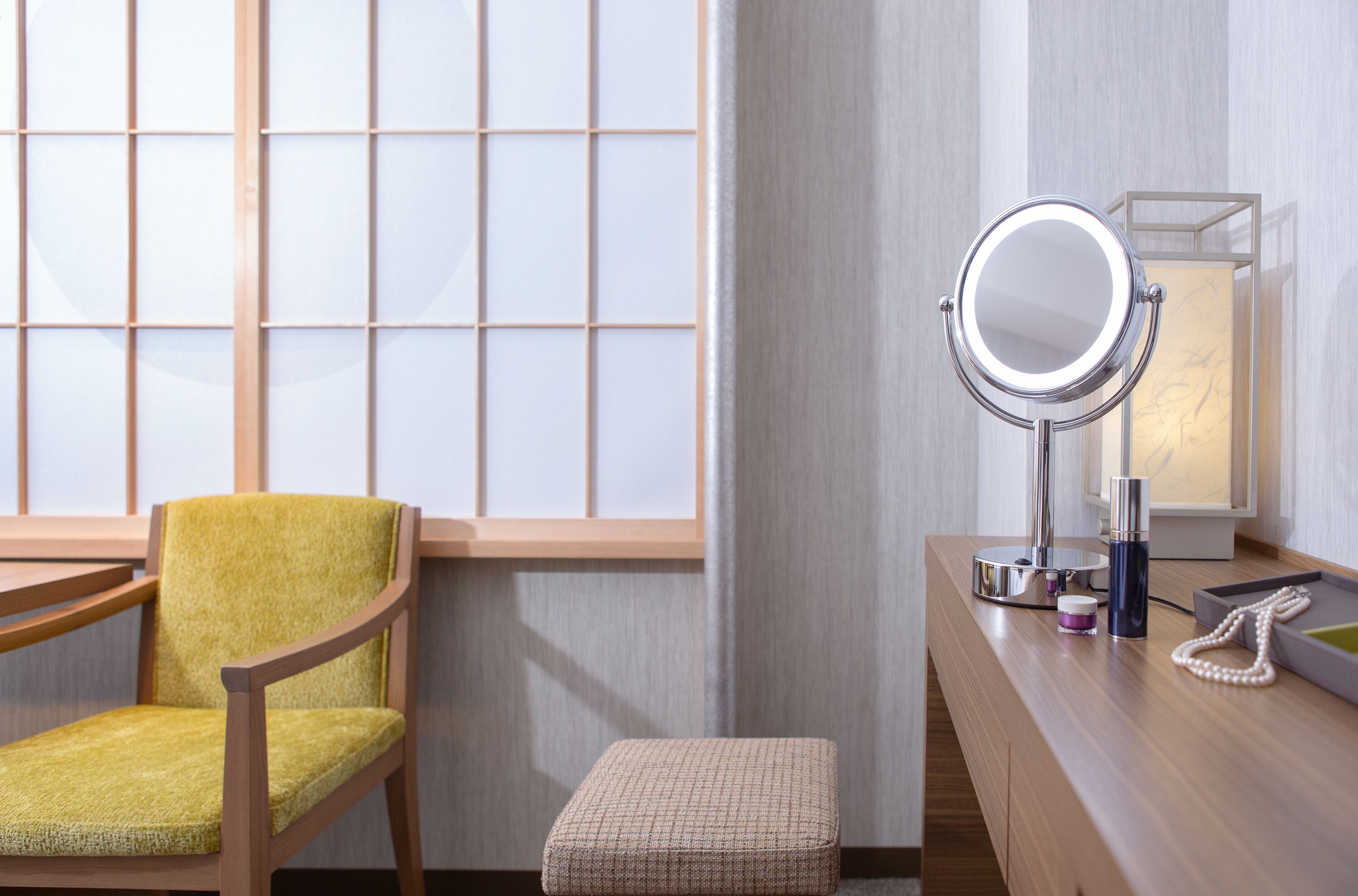 A room with a wooden chair, a small ottoman, a vanity with a mirror, a lamp, and a framed picture on the wall.