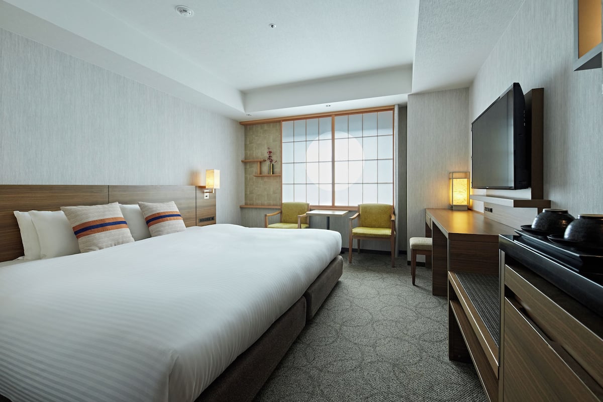 A hotel room with a large bed, a desk with a flat-screen TV, a chair, a small table, and a window with a view.