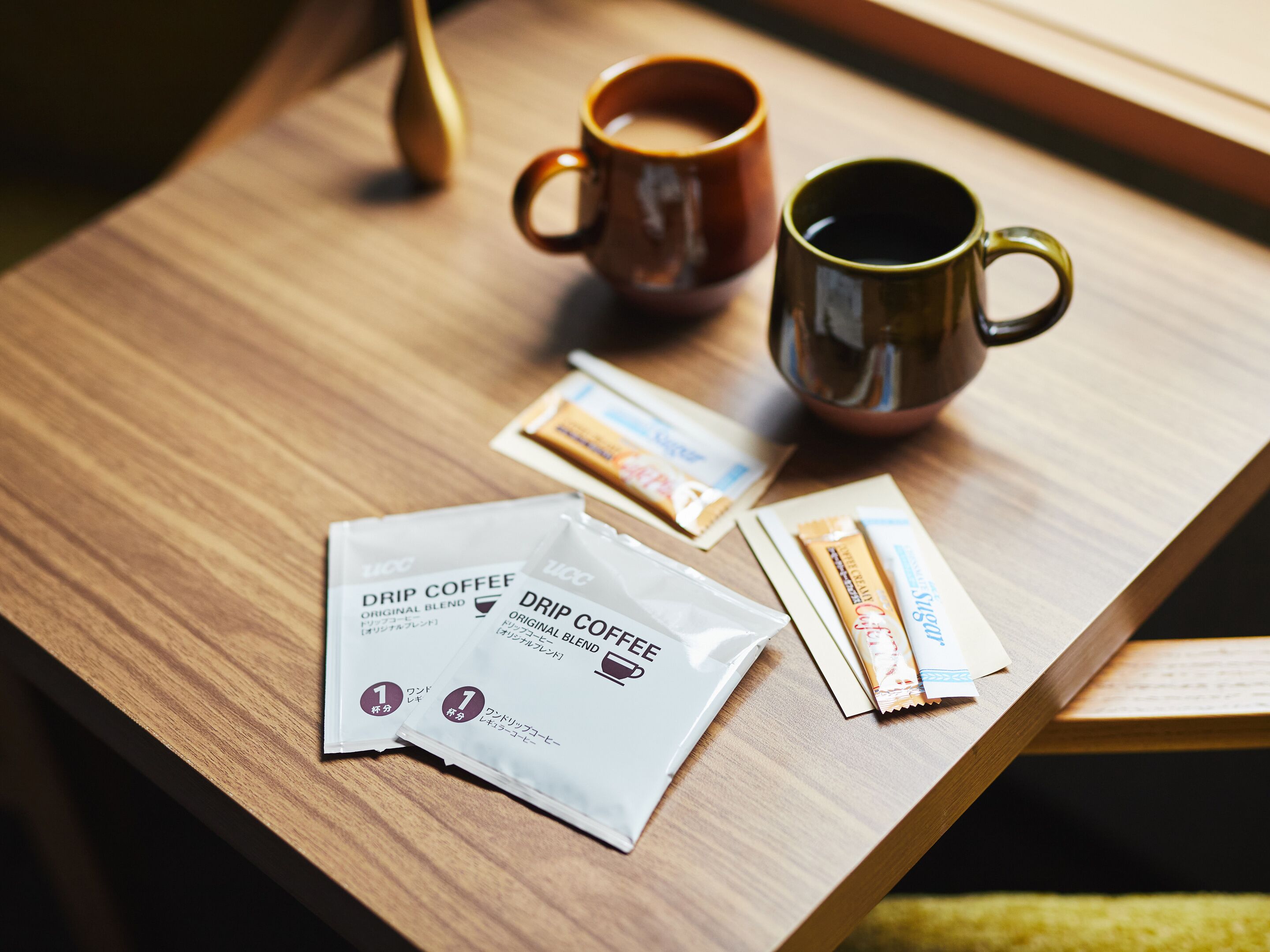 A wooden table with two coffee mugs, a drip coffee packet, and a packet of instant coffee.