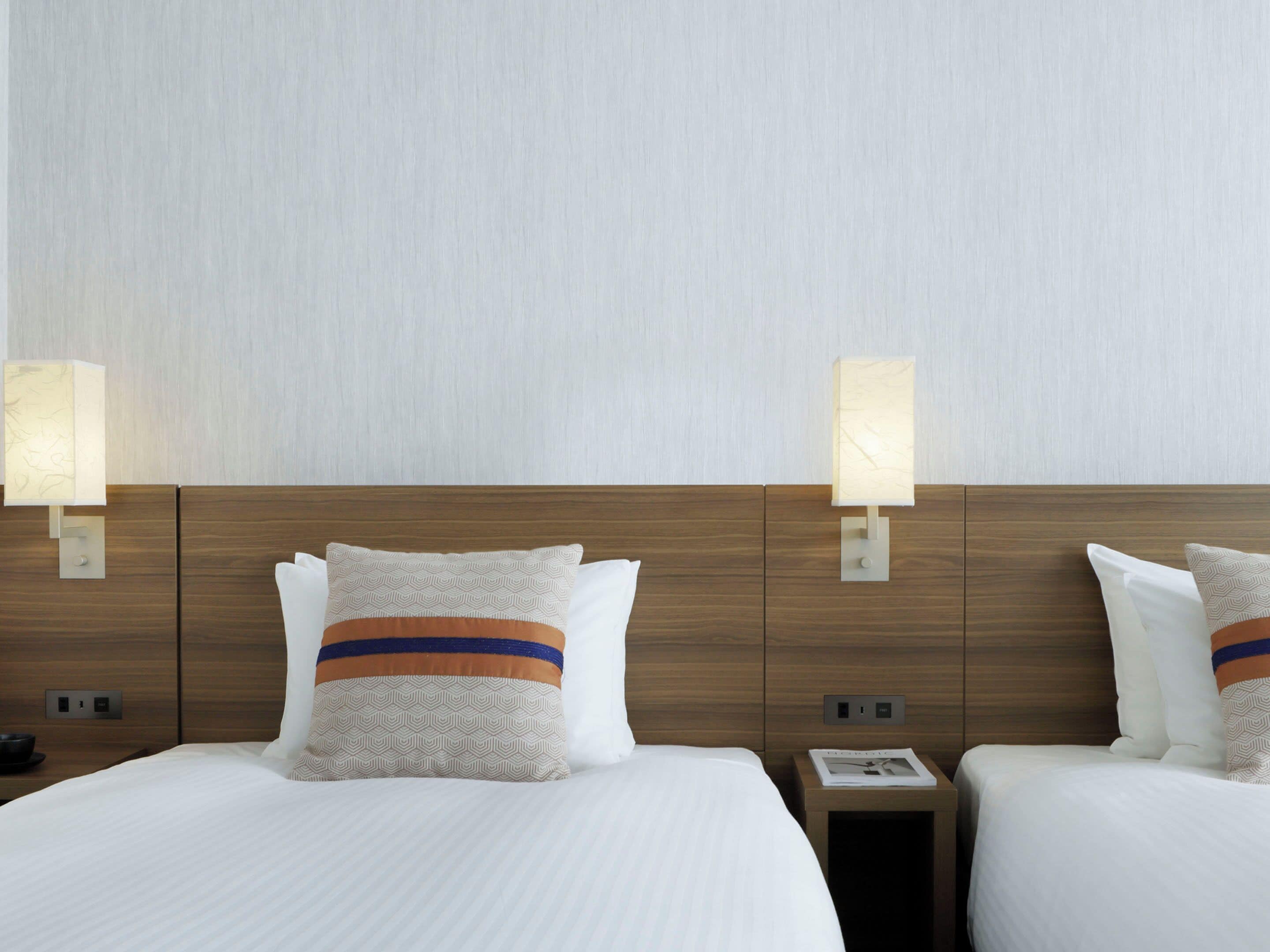 A hotel room with two beds, each with patterned pillows, bedside tables with lamps, and a plain wall.