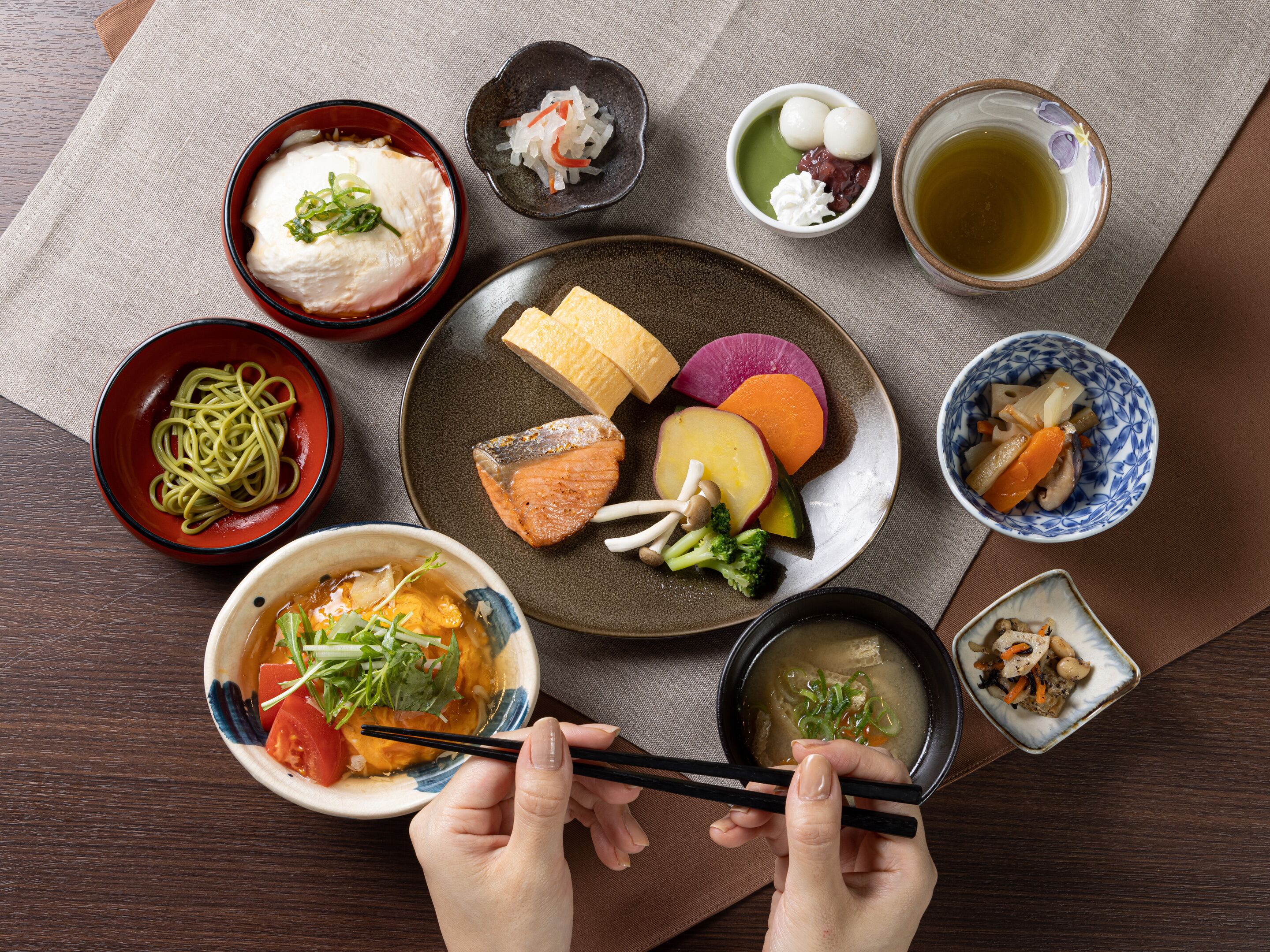 Diversos platos japoneses, como fideos, salmón y verduras, dispuestos en una mesa con palillos y una taza de té.