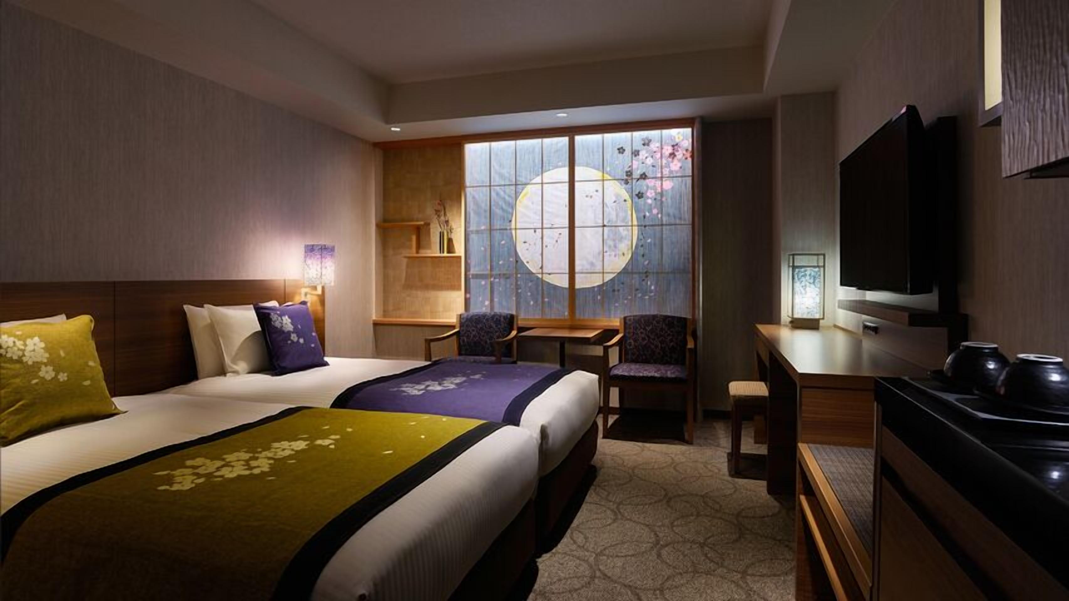 Quarto de hotel com duas camas, uma escrivaninha, televisão e uma janela grande com vista para flores de cerejeira.