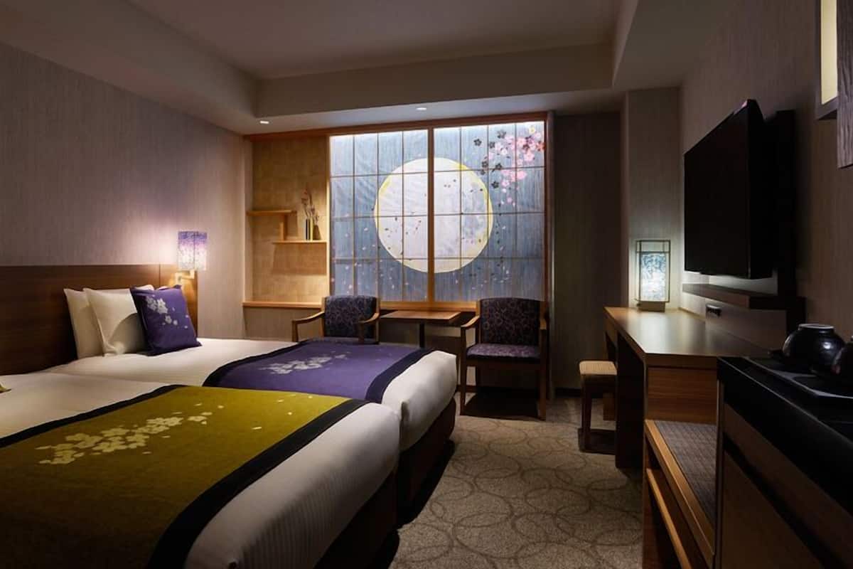 A hotel room with two beds, a desk, a TV, and a large window with a view of cherry blossoms.