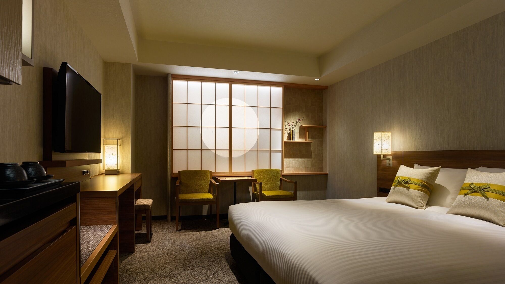 Quarto de hotel com uma cama, uma escrivaninha, duas cadeiras, televisão e janela com vista.