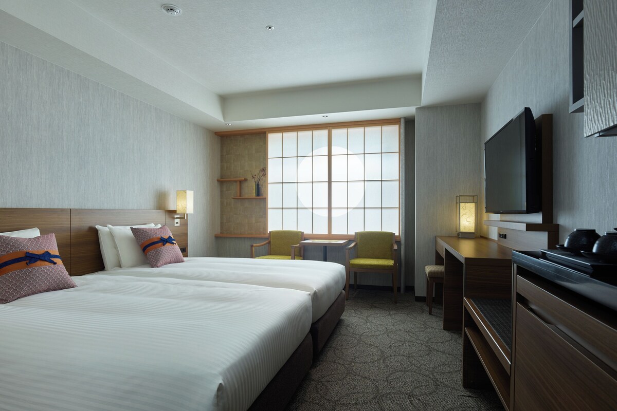A hotel room with two beds, a desk, a chair, a television, and a window with blinds.