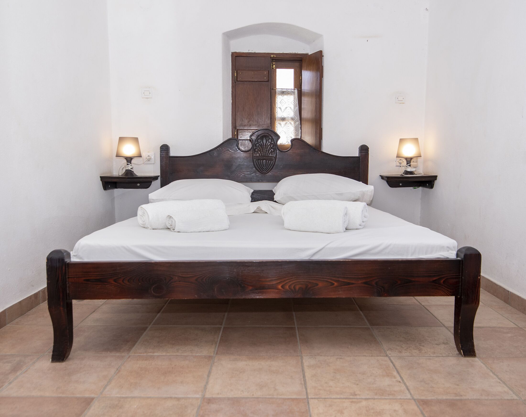 Een keurig opgemaakt bed met witte lakens en handdoeken, twee bedlampjes, een houten hoofdbord en een spiegel boven het bed.