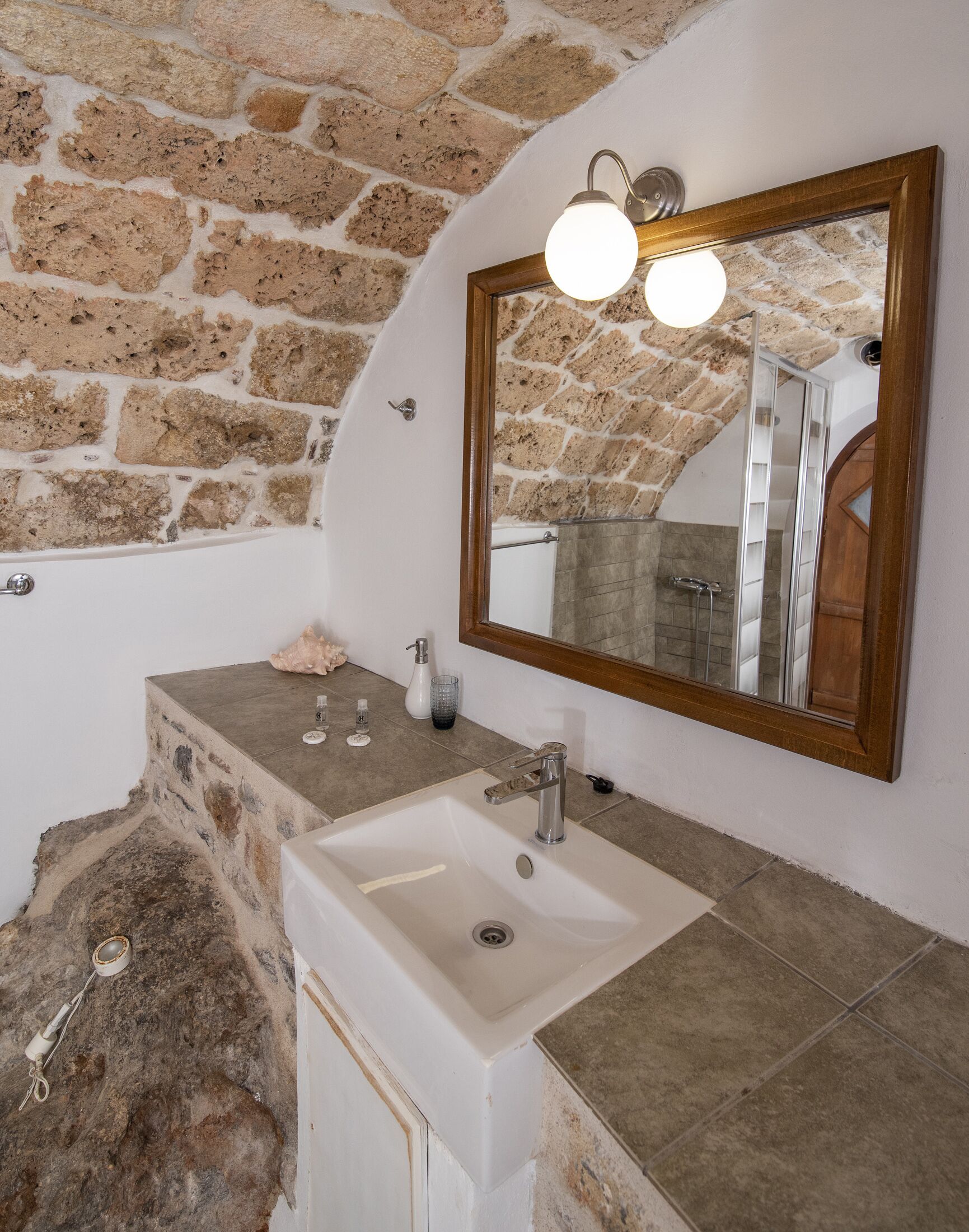 Un baño con pared de piedra, lavamanos blanco y espejo con marco de madera e iluminación.