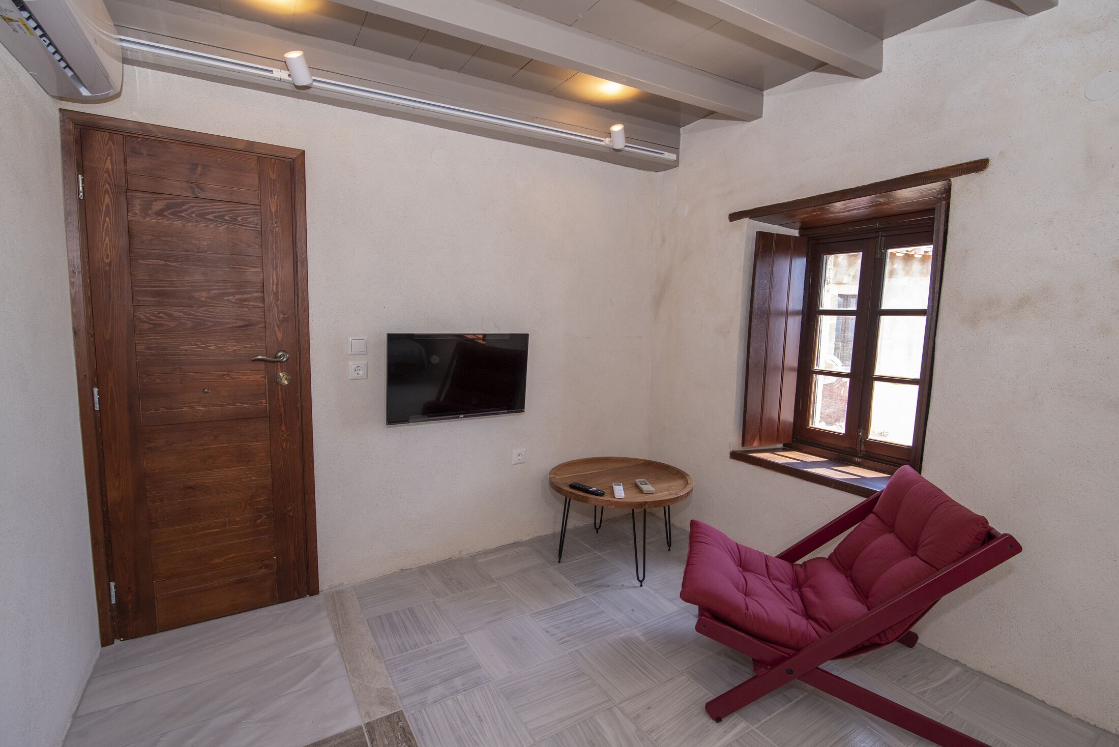 Una habitación con puerta de madera, un televisor de pantalla plana montado en la pared, una pequeña mesa redonda con un control remoto y una silla roja.