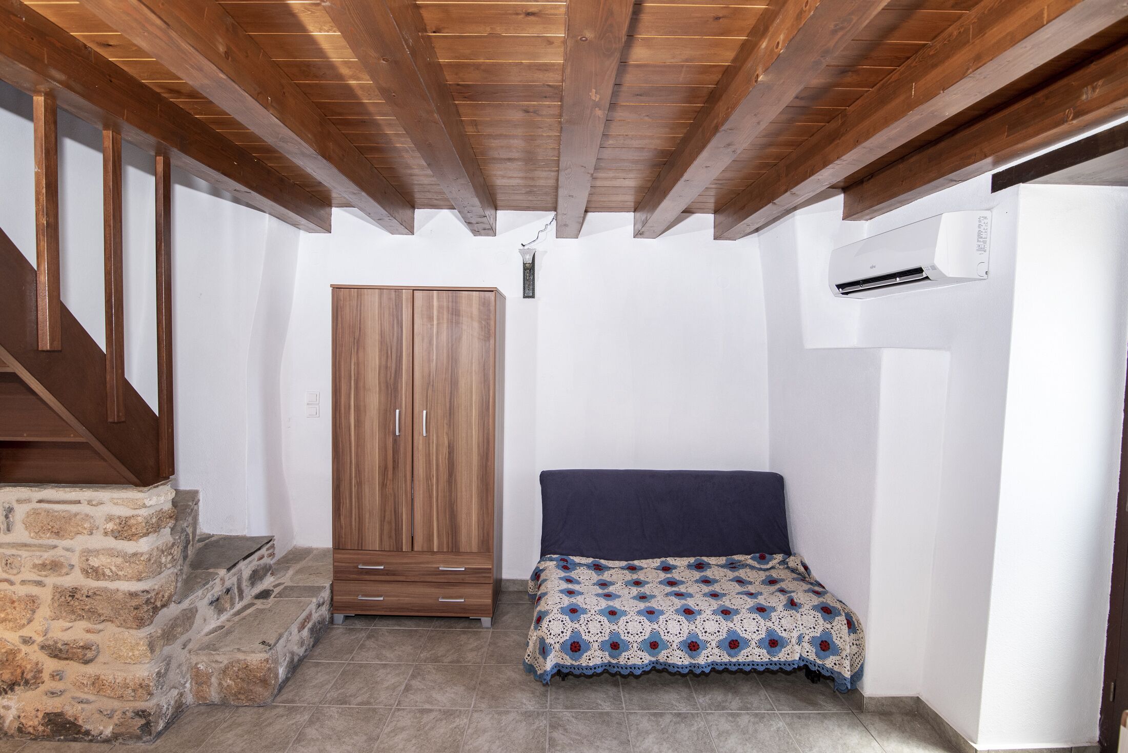 A small room with a wooden wardrobe, a bed with a patterned bedspread, and a stone staircase.
