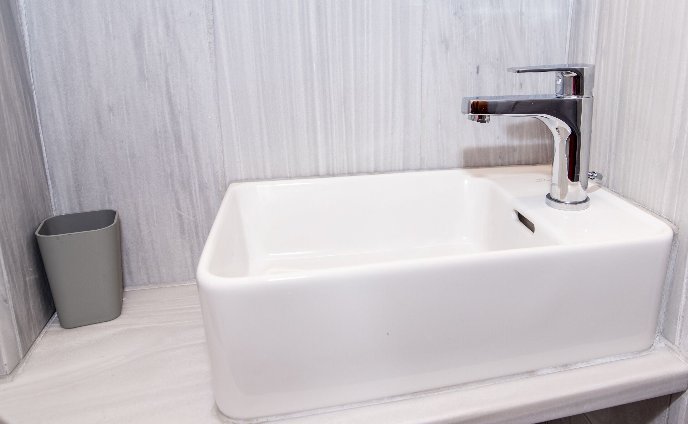 A white bathroom sink with a chrome faucet, a small trash bin, and a tiled wall.