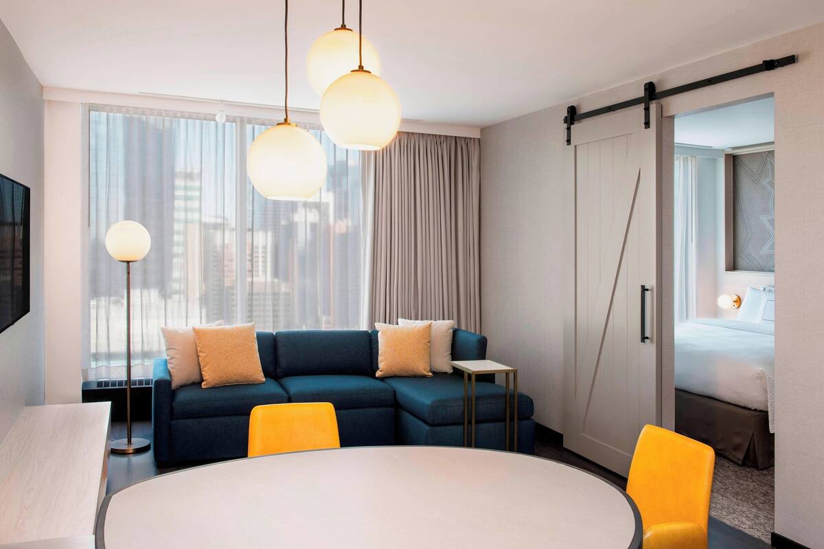 A modern hotel room with a sectional sofa, a dining table, a small side table, and a bed with a view.