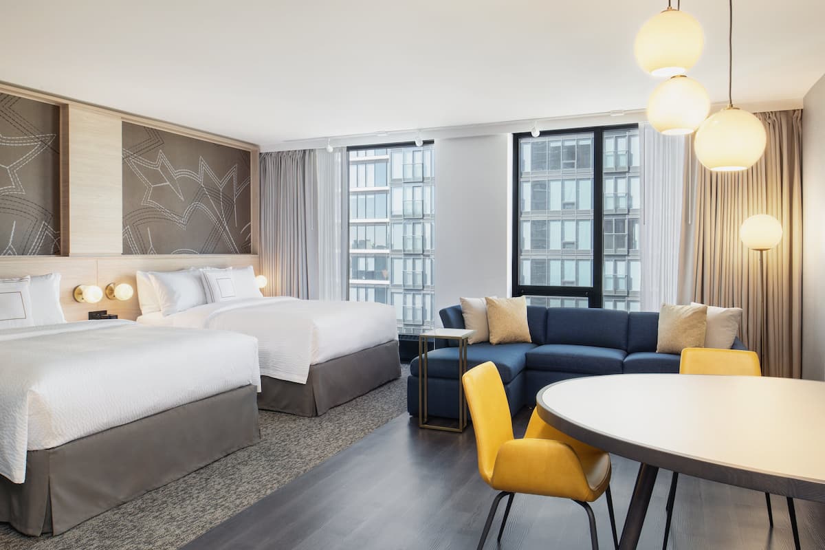 A hotel room with two beds, a sofa, a dining table, and large windows.