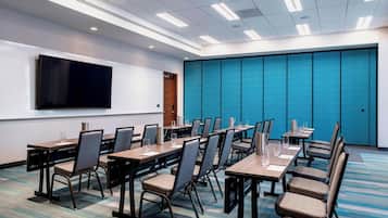 A conference room with multiple tables and chairs, a large flat-screen TV mounted on the wall, and a blue partition wall.