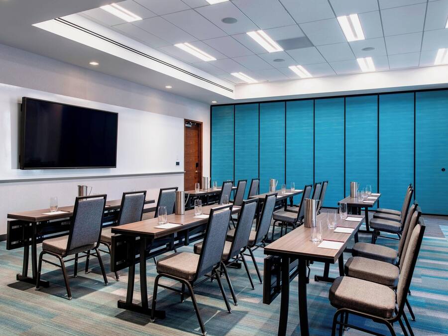 A conference room with multiple tables and chairs, a large flat-screen TV mounted on the wall, and a blue partition wall.