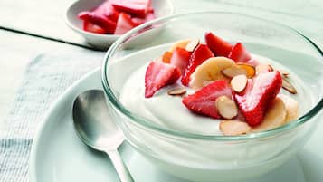 Un bol de yaourt garni de fraises et d’amandes, posé sur une assiette blanche, avec une cuillère à côté.