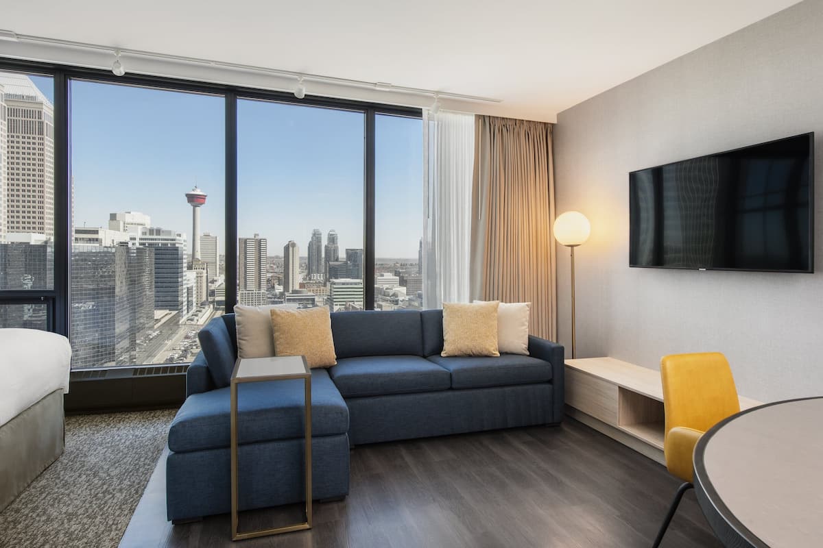 A modern hotel room with a large window offering a city view, a sofa, a bedside table, a lamp, and a TV on the wall.