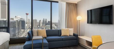 A modern hotel room with a large window offering a city view, a sofa, a bedside table, a lamp, and a TV on the wall.