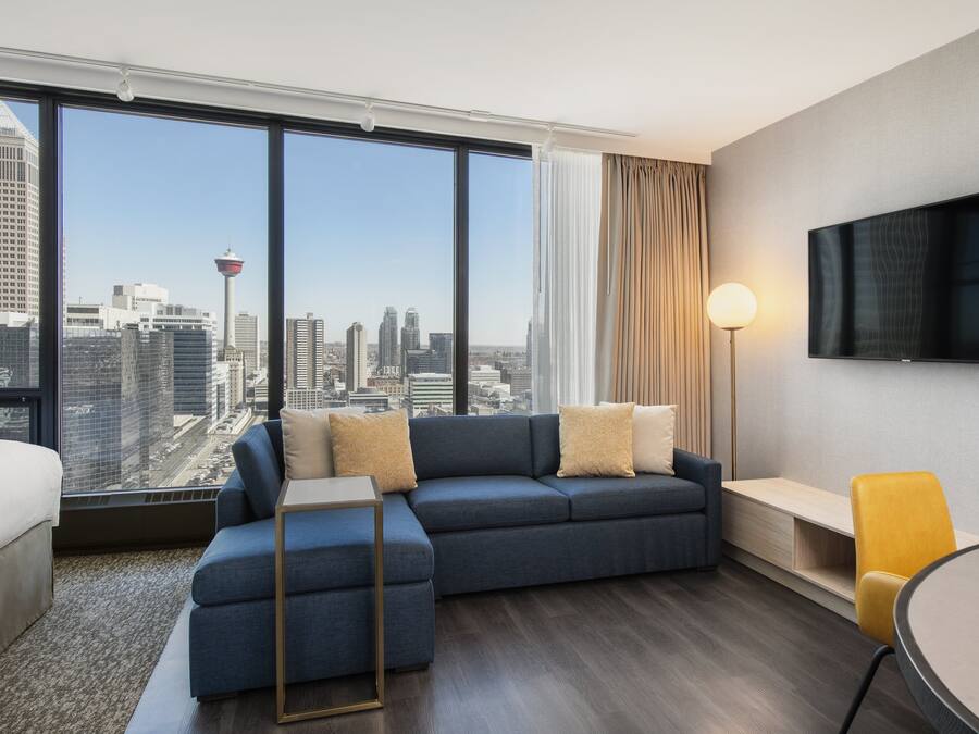 A modern hotel room with a large window offering a city view, a sofa, a bedside table, a lamp, and a TV on the wall.