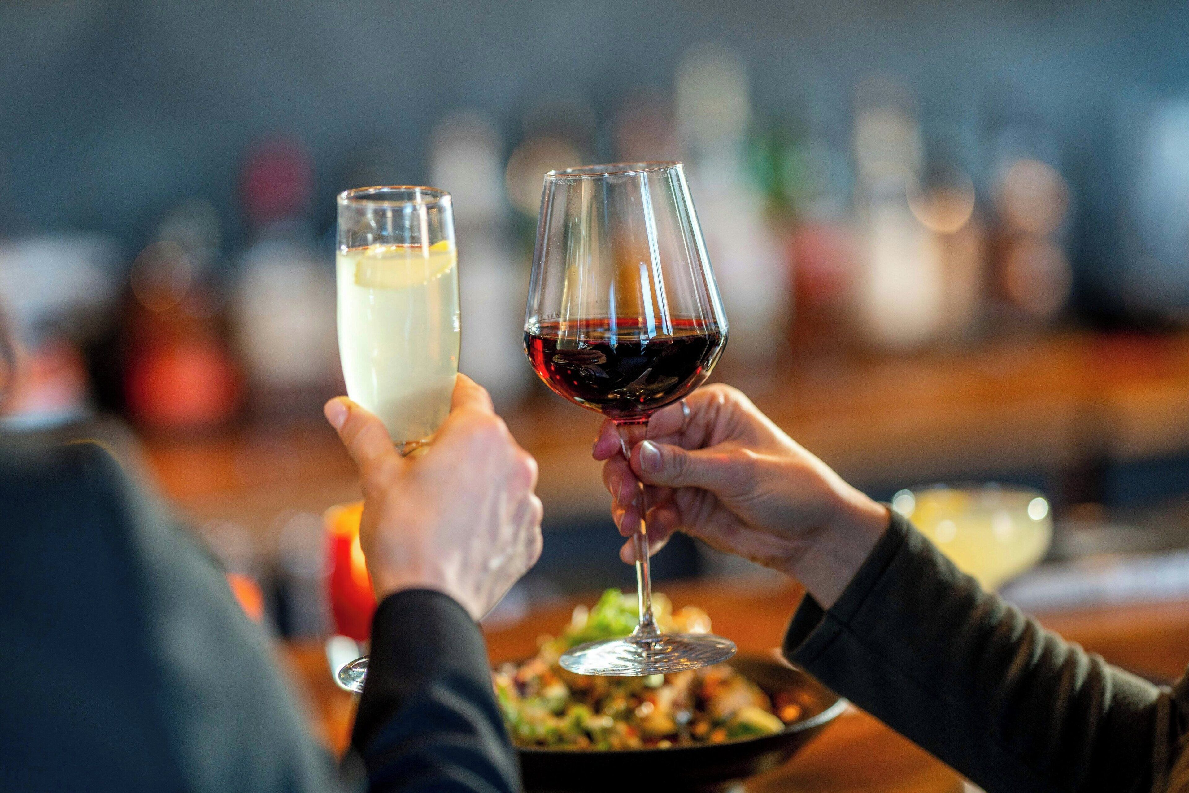 Dos manos sosteniendo copas de vino, una con vino blanco y la otra con vino tinto, en un bar con fondo desenfocado.