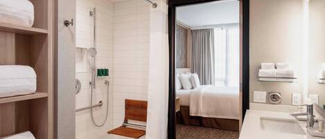 A hotel bathroom with a shower, a bench, a sink, and a bed visible through an open door.