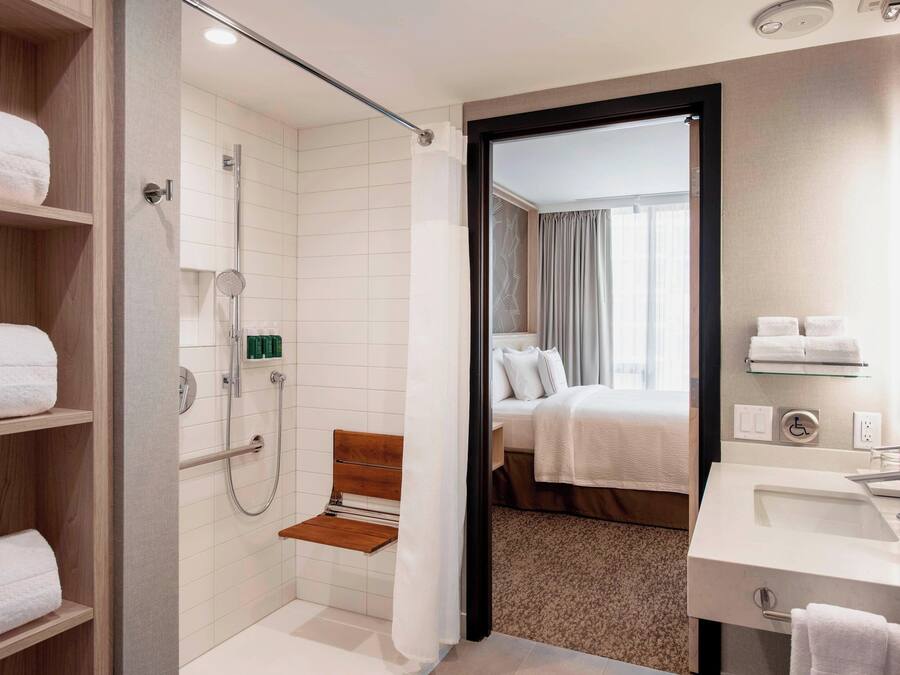 A hotel bathroom with a shower, a bench, a sink, and a bed visible through an open door.