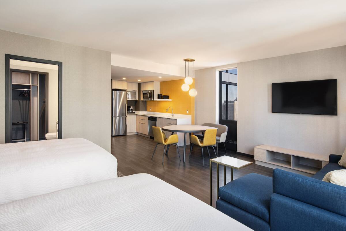 A modern hotel room with a large bed, a sofa, a dining area with yellow chairs, a kitchen area, and a TV mounted on the wall.
