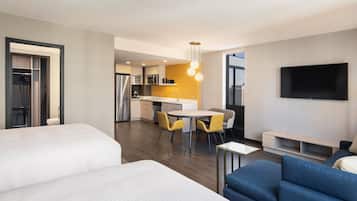 A modern hotel room with a large bed, a sofa, a dining area with yellow chairs, a kitchen area, and a TV mounted on the wall.