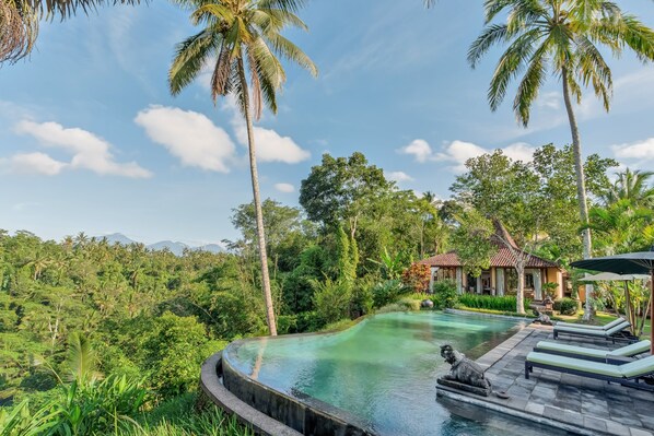 A pool with a curved edge, surrounded by lush greenery and palm trees, with a traditional building in the background.