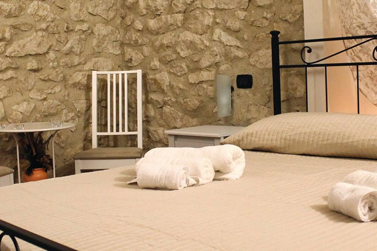 Un lit bien fait, avec des serviettes pliées, un mur en pierre, une chaise blanche et une petite table sur laquelle est posé un vase.
