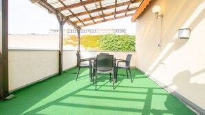 Eine überdachte Terrasse mit Tisch und Stühlen, einem grünen Kunstrasen und Blick auf ein Gebäude und Bäume im Hintergrund.