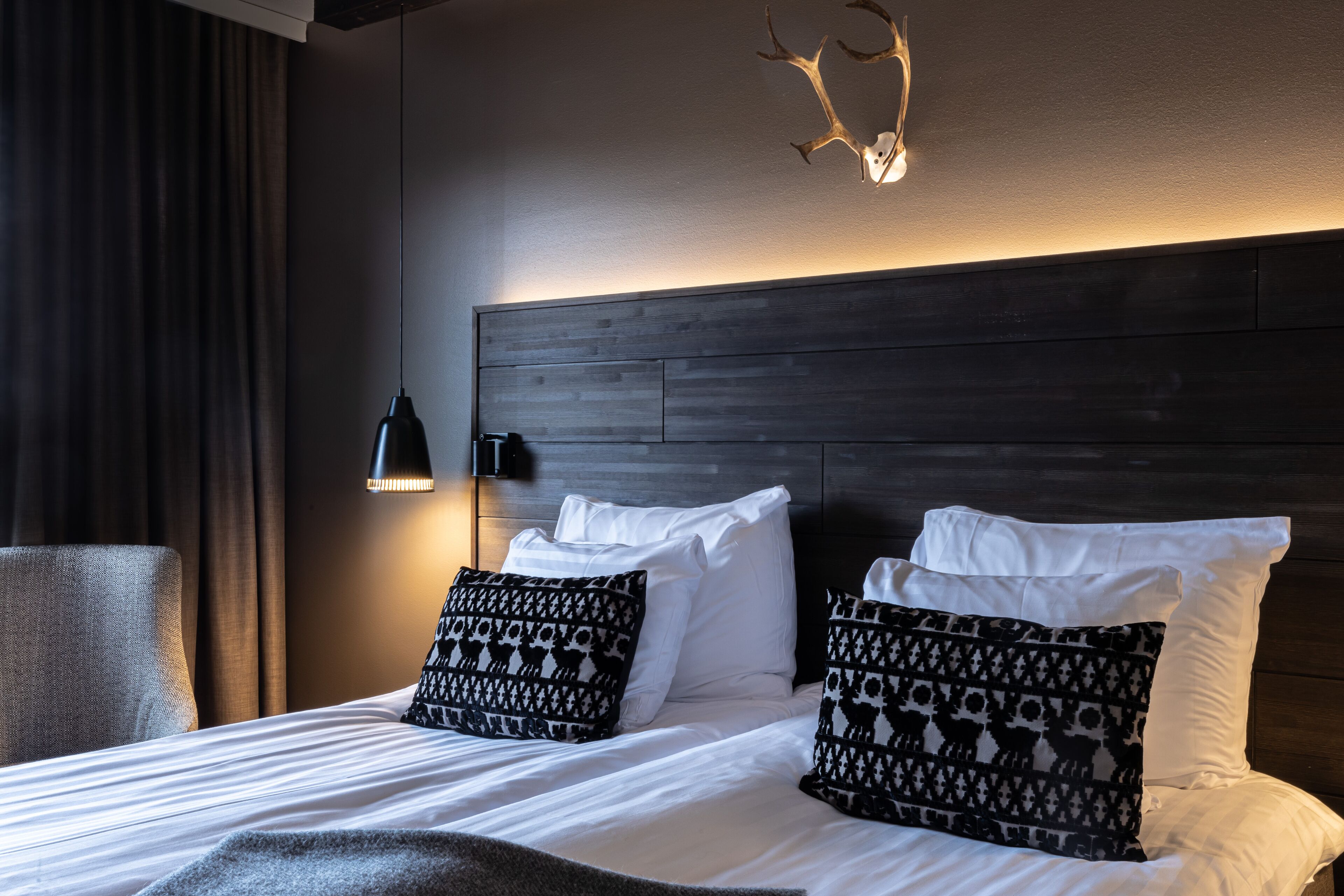 Un lit bien fait, avec du linge de lit blanc et des coussins à motifs, une tête de lit en bois foncé, un luminaire fixé au mur et un rideau sur le côté gauche.