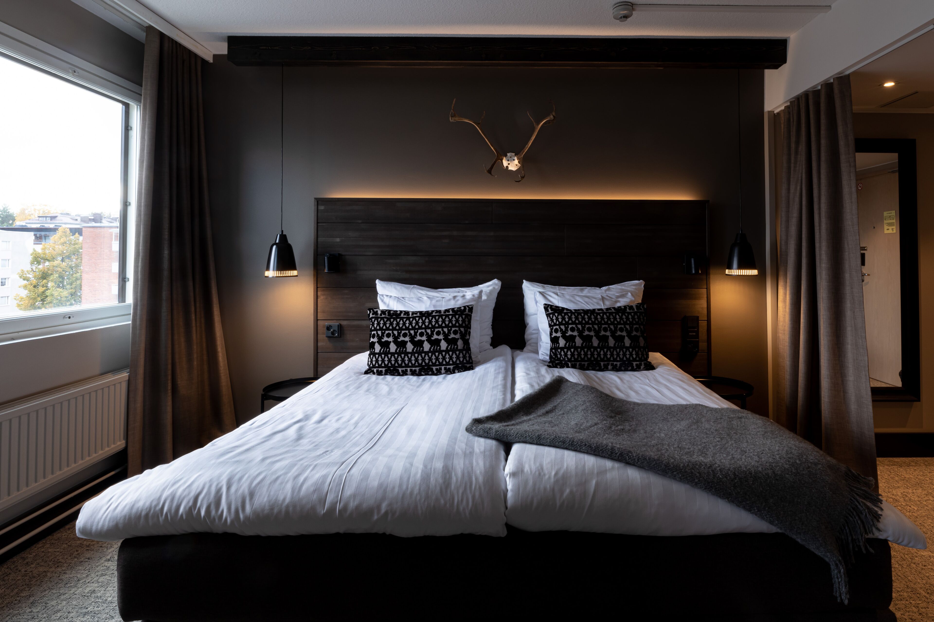 Habitación de hotel moderna con una cama grande, dos lámparas de noche, un adorno de asta de ciervo en la pared y vistas de la ciudad a través de la ventana.