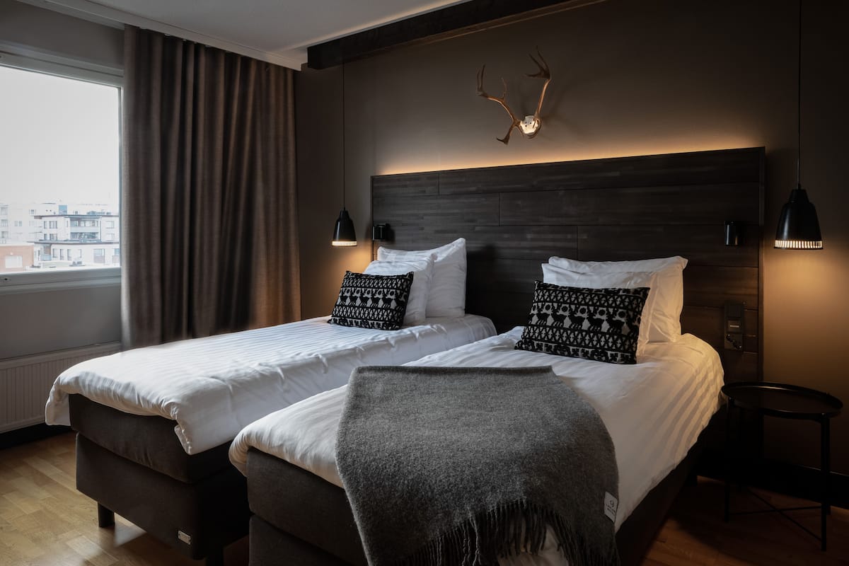 A hotel room with two beds, a large window with curtains, a wooden headboard, and a mounted antler decoration.
