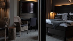 A modern hotel room with a large bed, a flat-screen TV mounted on the wall, a comfortable seating area with a chair and a small table, and a large mirror reflecting the room. - Lapland Hotels Kuopio (Kuopio)