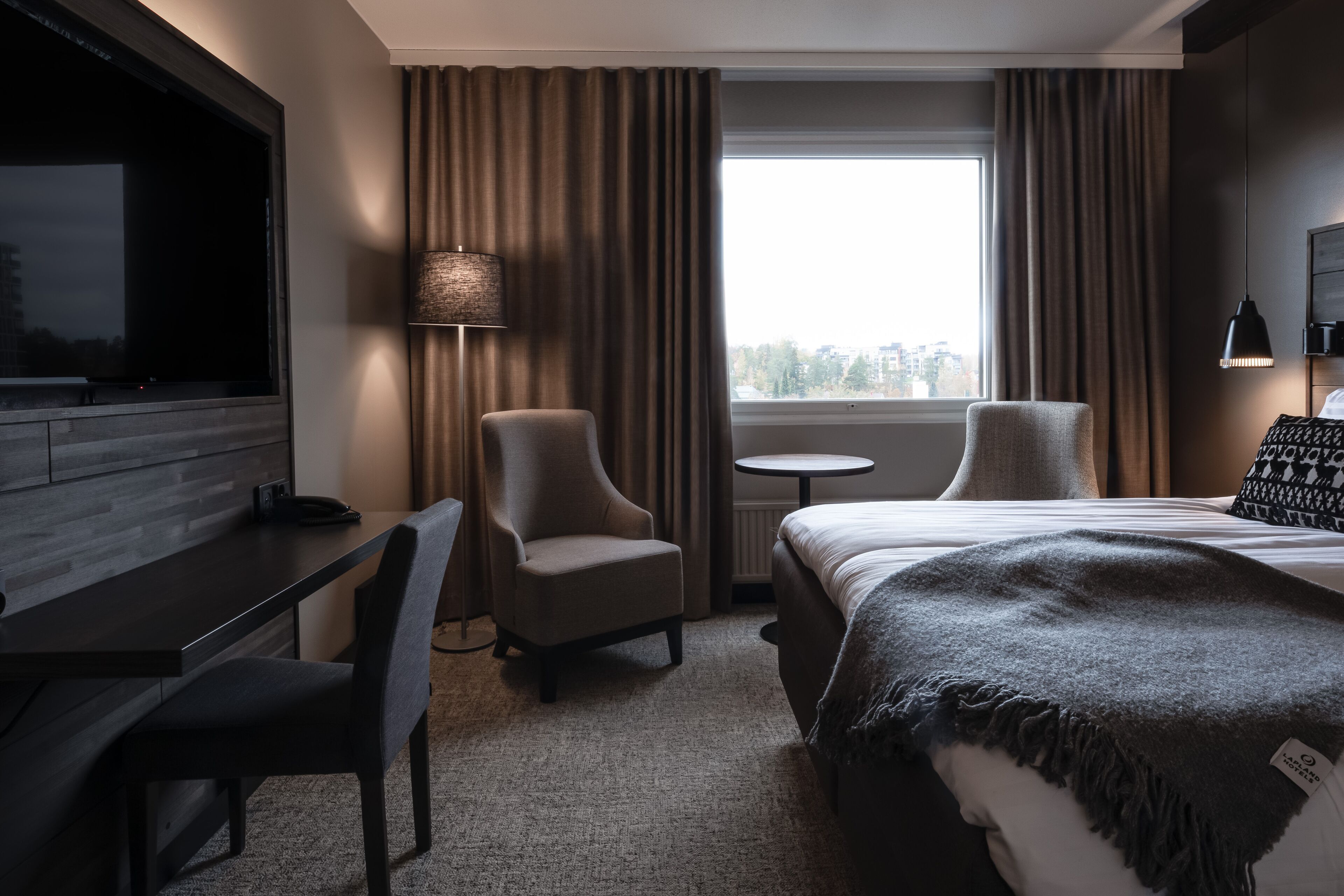 A hotel room with a bed, two armchairs, a desk, a lamp, a small table, and a TV.