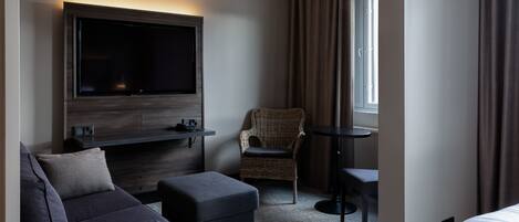 A hotel room with a flat-screen TV mounted on the wall, a sofa, an ottoman, a wicker chair, and a small table.