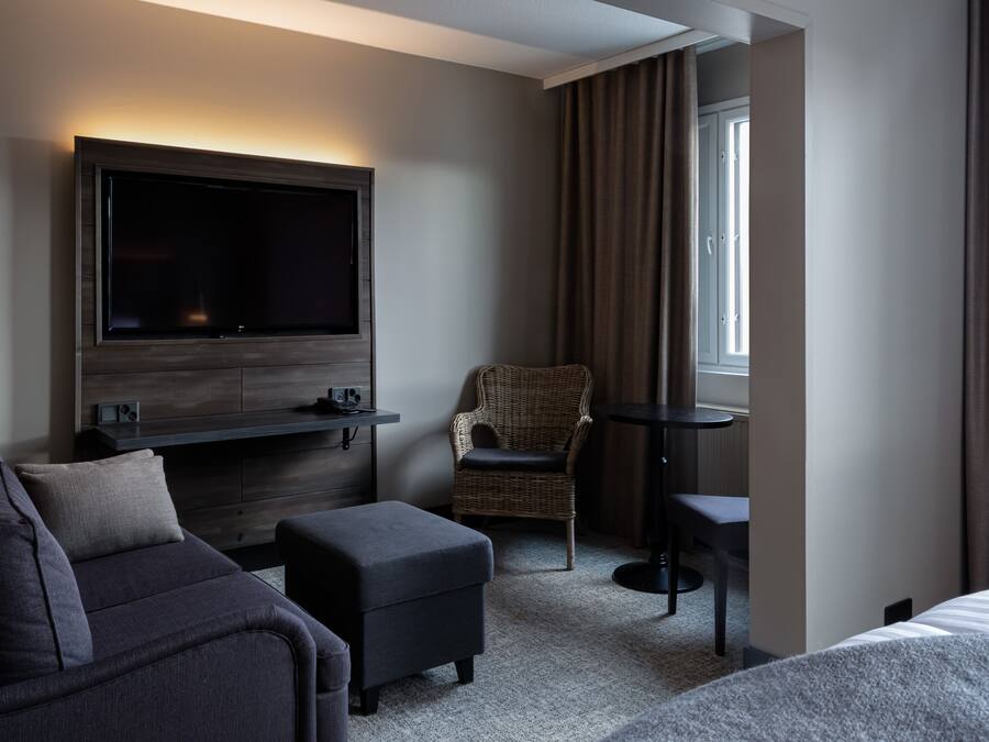 A hotel room with a flat-screen TV mounted on the wall, a sofa, an ottoman, a wicker chair, and a small table.