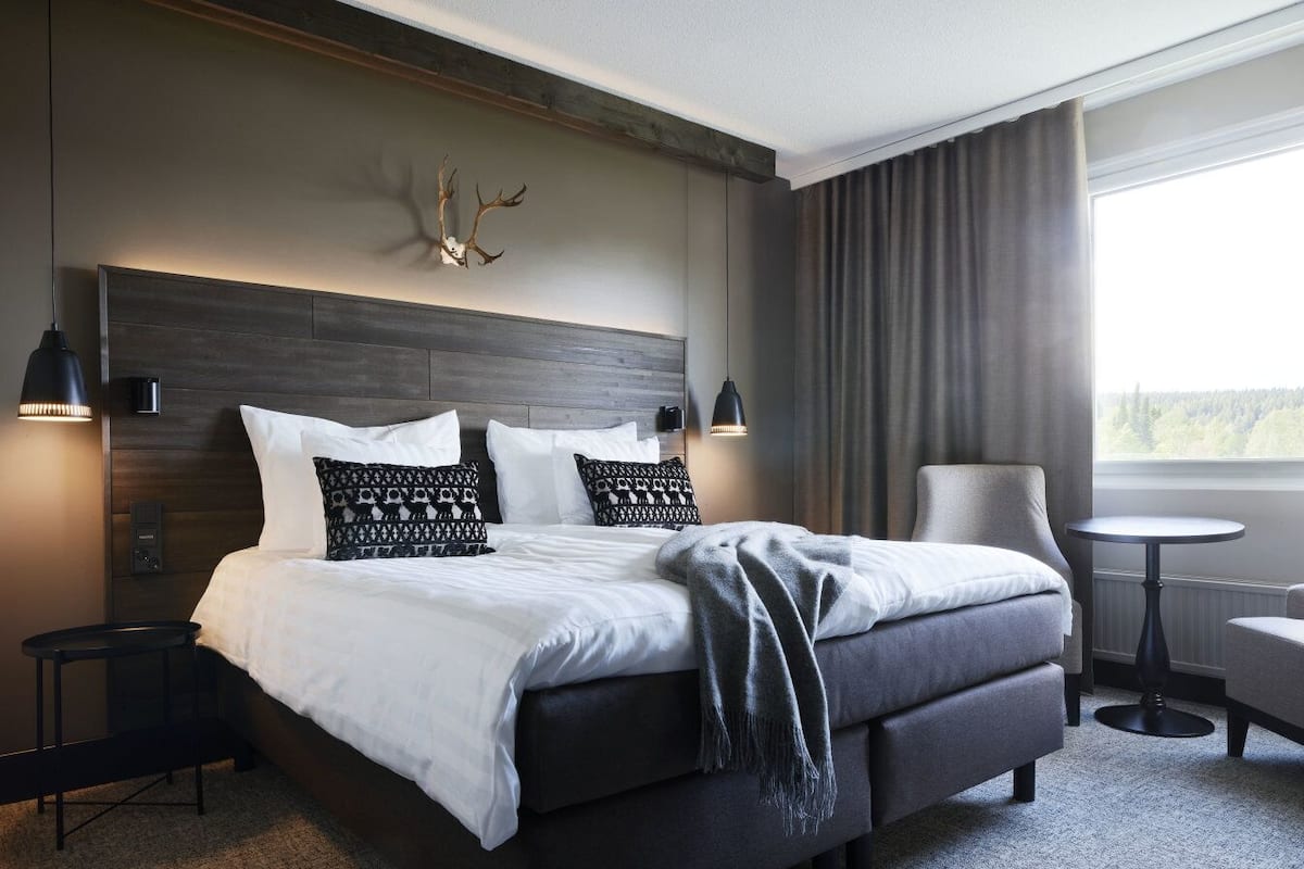 A modern hotel room with a large bed, two bedside tables, a chair, a small round table, and a large window with curtains.