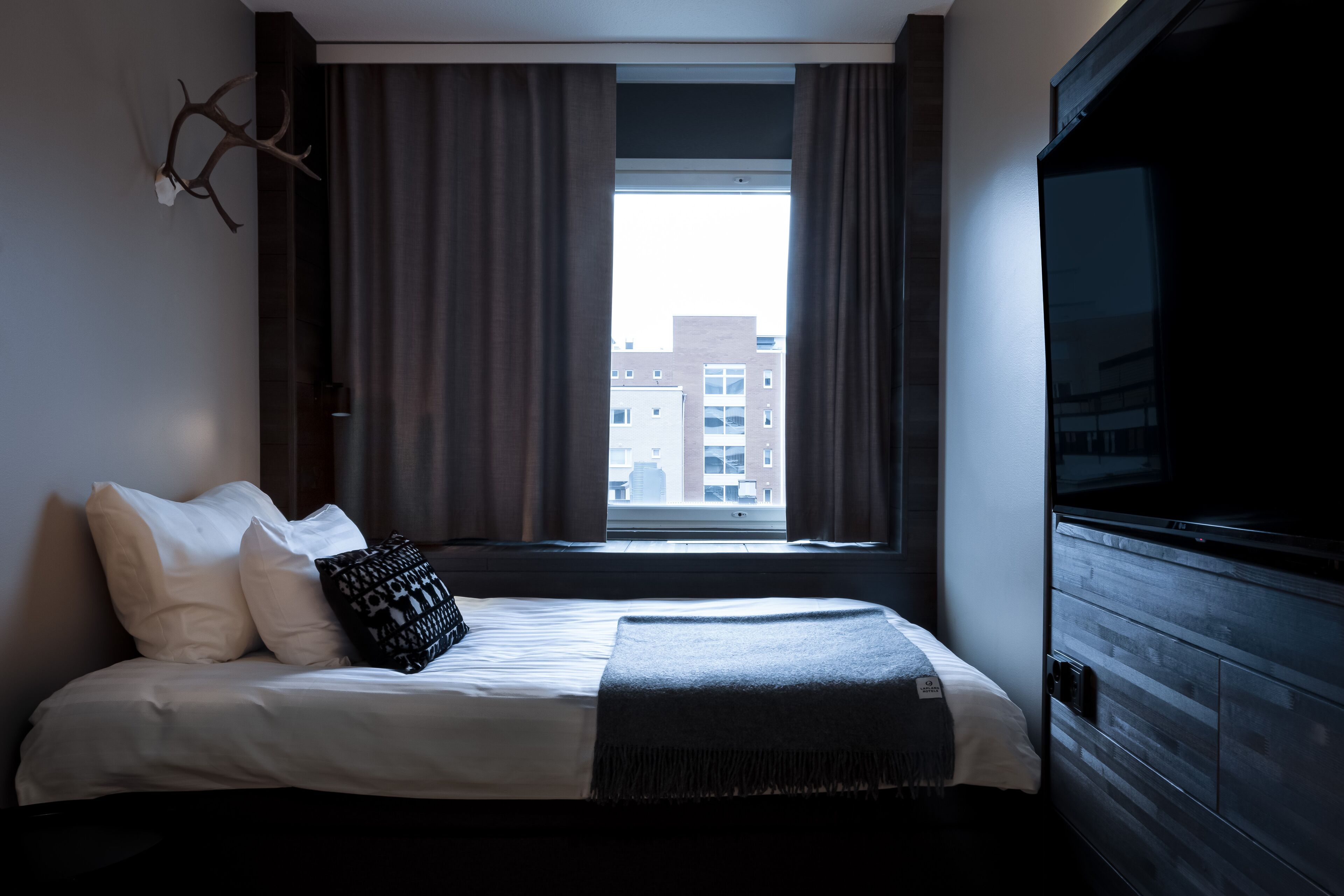 A modern hotel room with a large bed, a city view through the window, and a television on the right.
