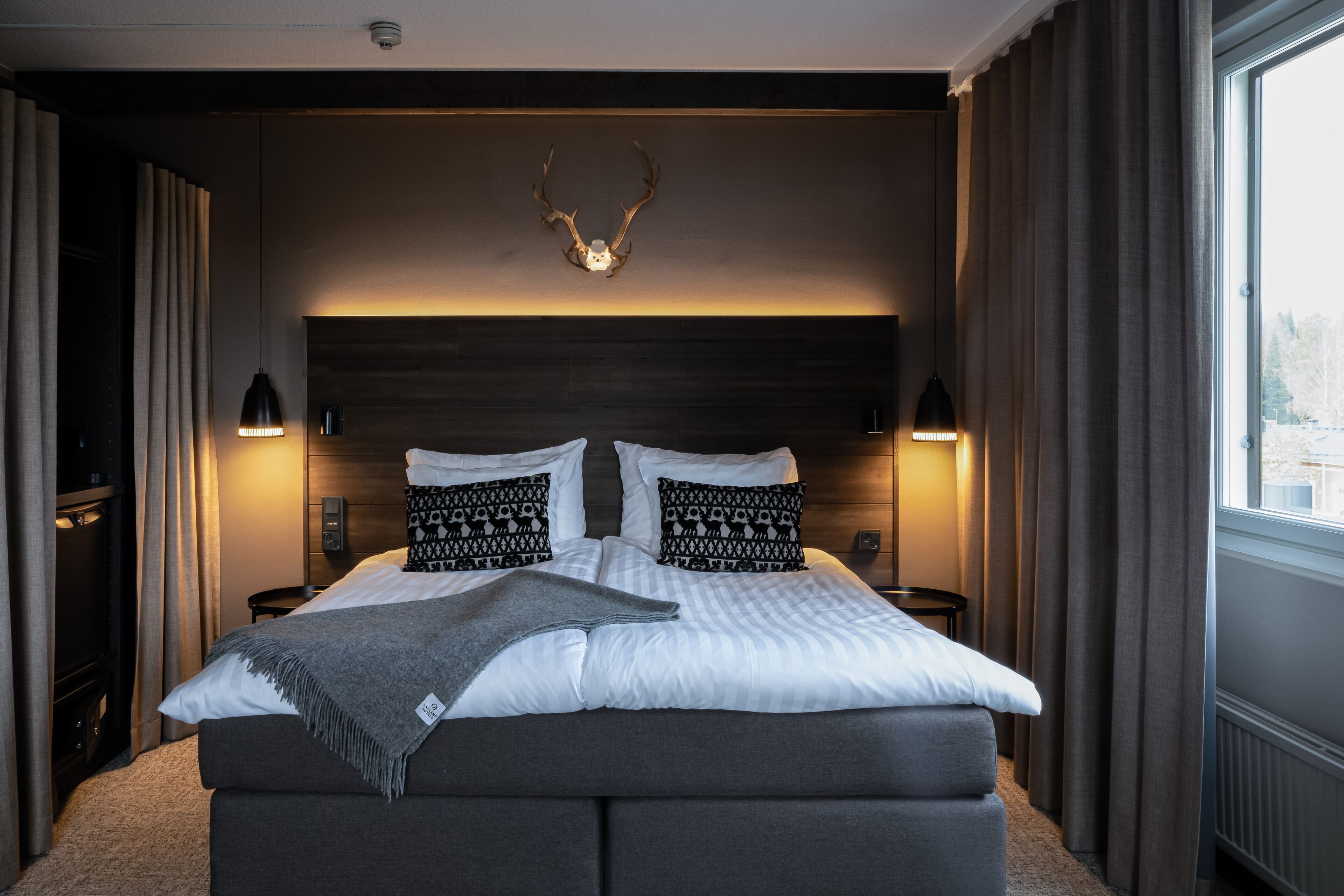 A bedroom with a large bed, two bedside lamps, a headboard with antlers, and a window with curtains.