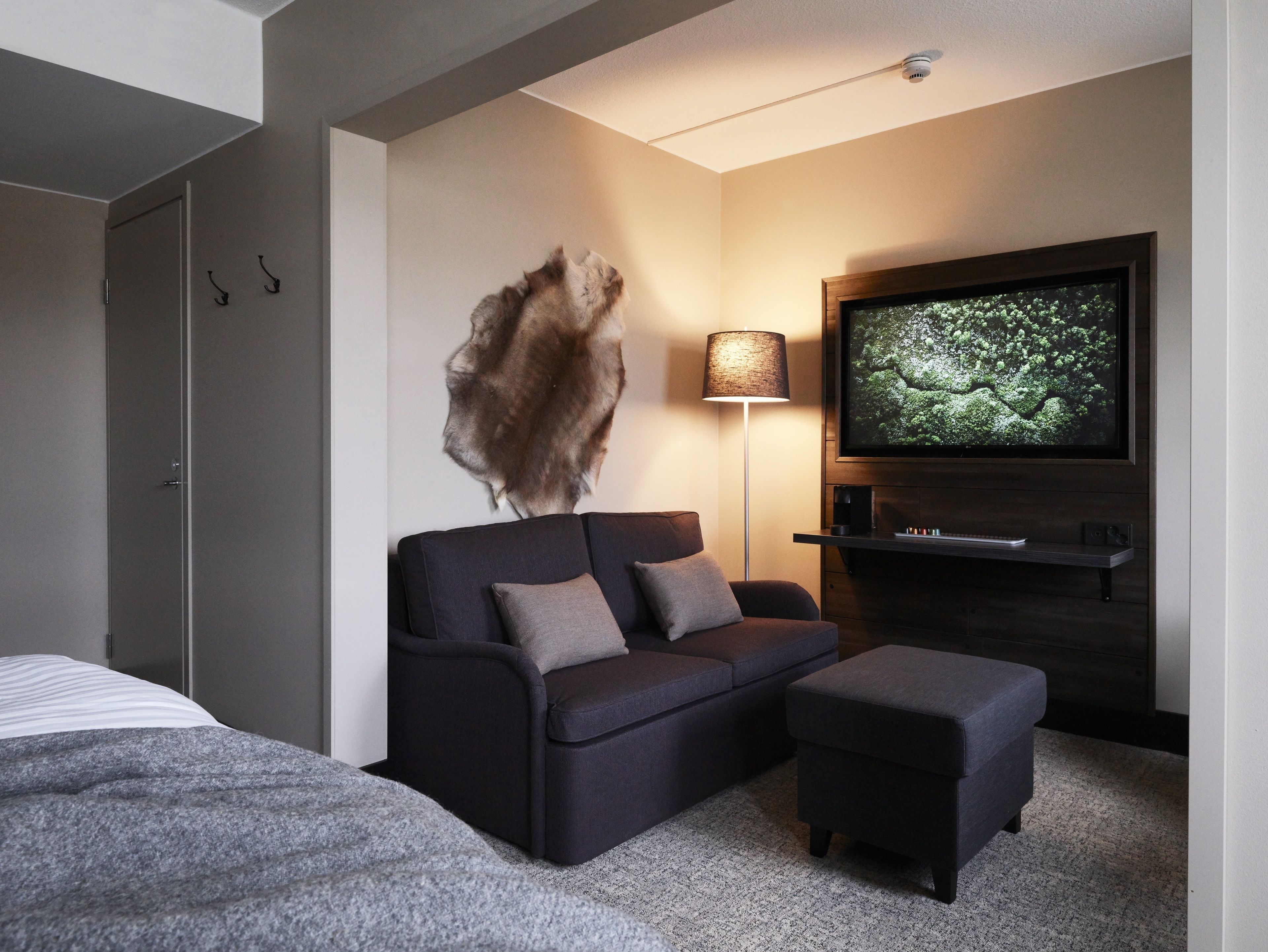Une chambre d’hôtel moderne équipée d’un canapé, d’un pouf et d’un téléviseur à écran plat fixé au mur.