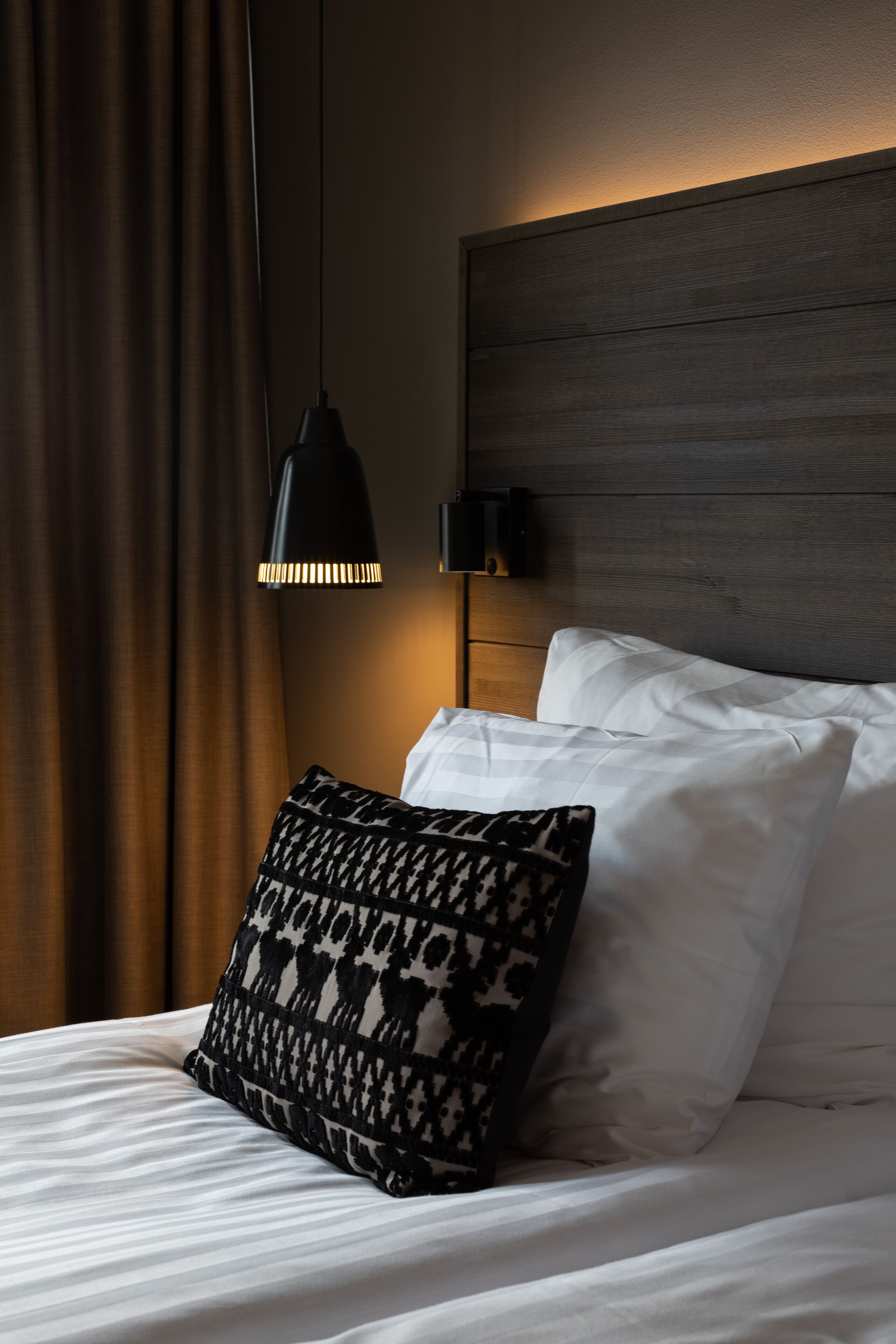 Una cama bien hecha con sábanas blancas y un cojín estampado, un cabecero de madera oscura y una lámpara colgante negra.
