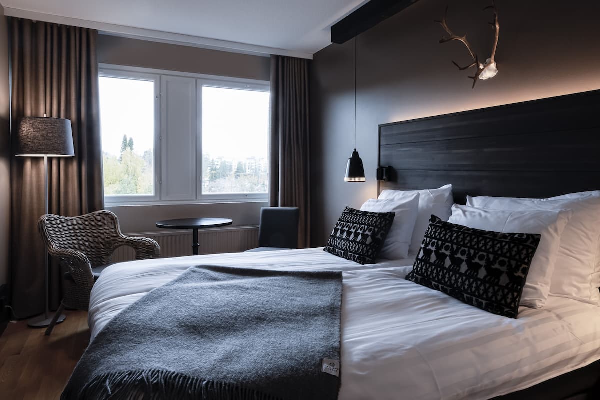 A modern hotel room with a large bed, a small round table, a chair, and a view of the city through large windows.
