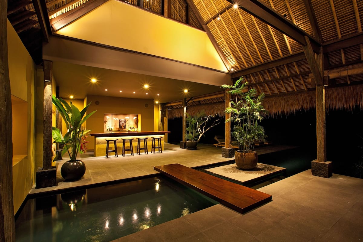 A well-lit indoor pool area with a bar and seating, surrounded by potted plants and a thatched roof structure.
