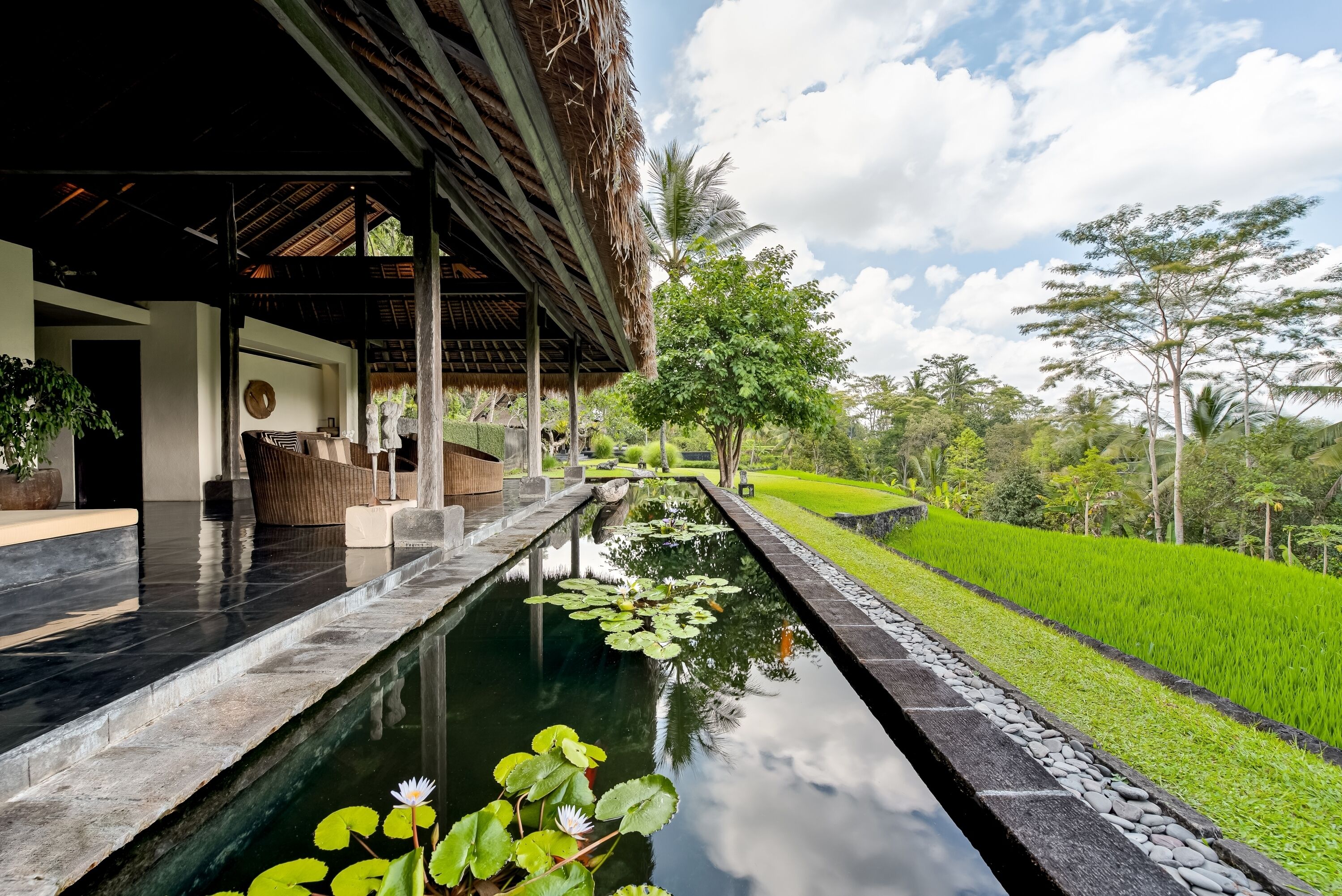 A serene outdoor pool area with a thatched roof structure, surrounded by lush greenery and a well-maintained garden.